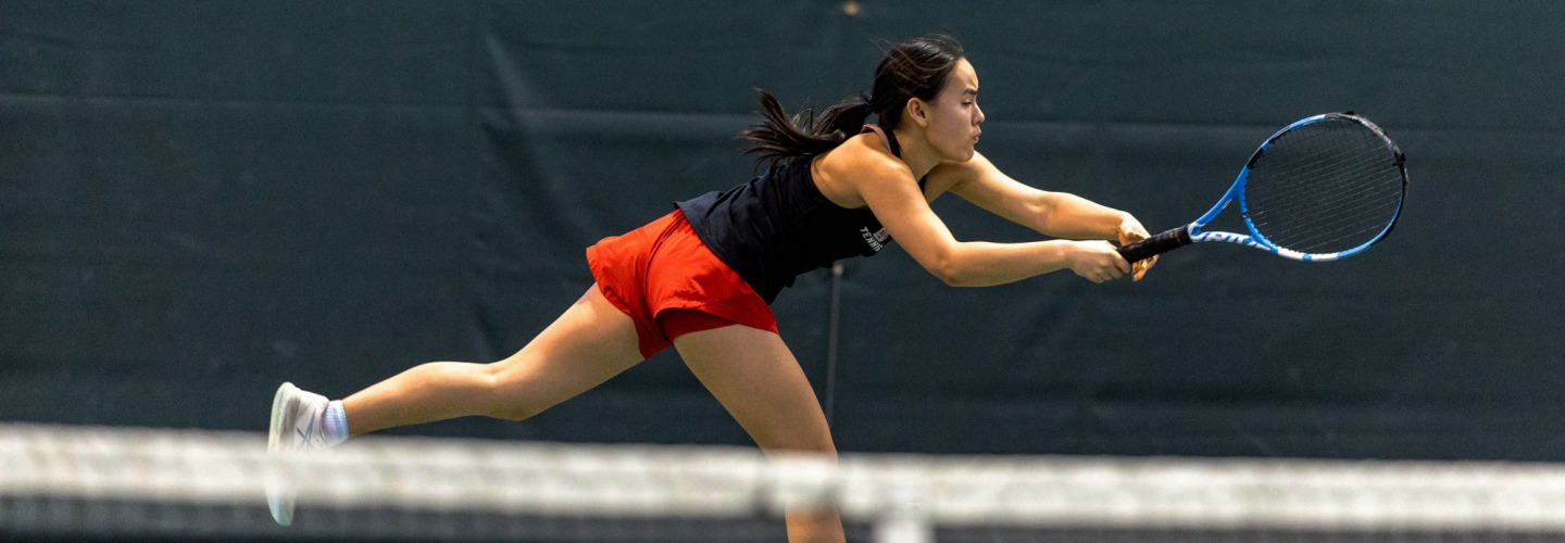 Women's tennis player in black top and scarlet shorts stretches out with her tennis racket 