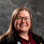 White, cis-gender woman with long hair and glasses, burgundy shirt, black jacket, and owl pin