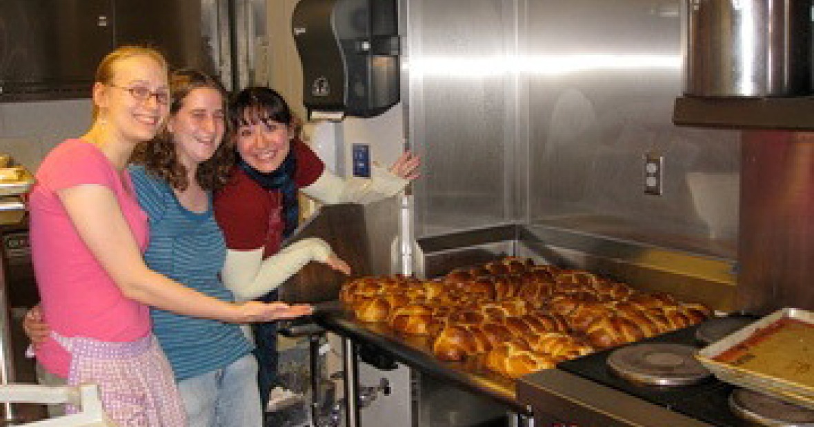 Three women in an industrial style kitchen stand to the left of and gesture toward a tray of baked challah loaves