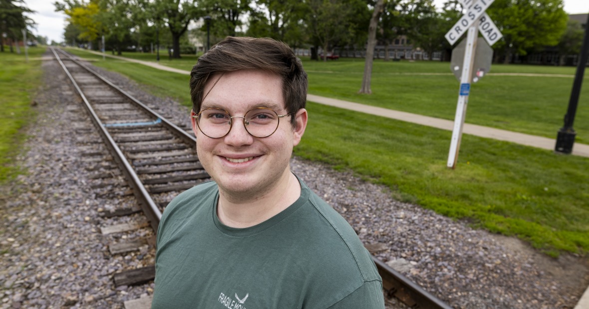 A man with short brown hair smiles in front of train tracks. He wears brown glasses and a teal shirt.