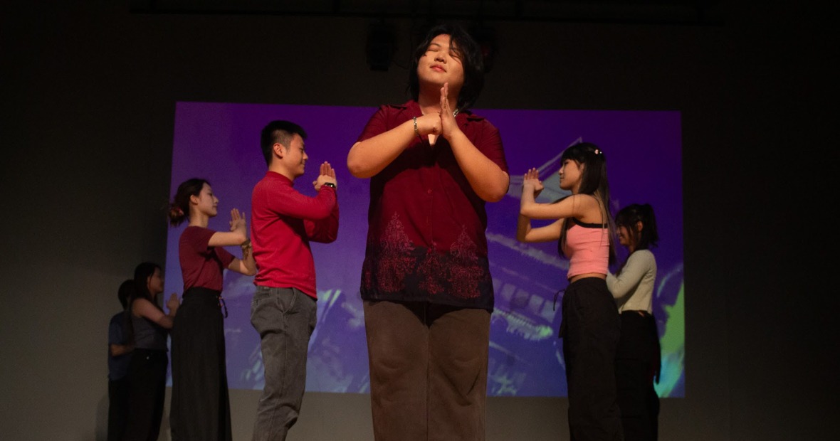 Five people stand on a stage. Four of the five face each other and hold their hands together, while the fifth person stands facing the viewer, smiling, and creates a fist held up against a flat palm with their hands
