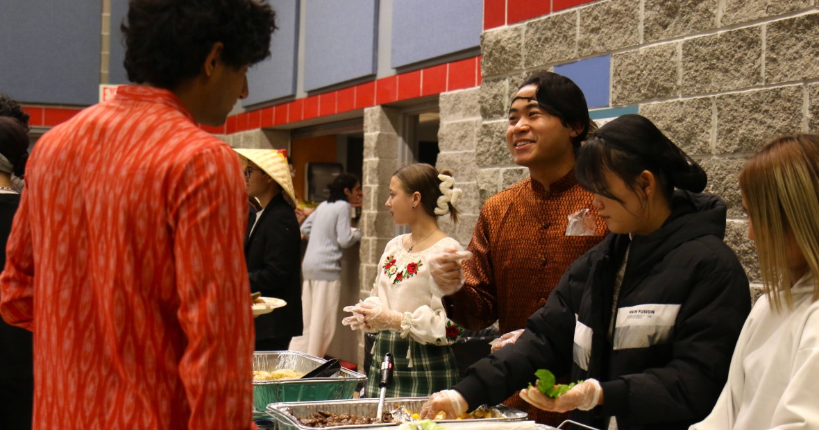 Students wearing hairnets and gloves stand in front of multiple tins of food 