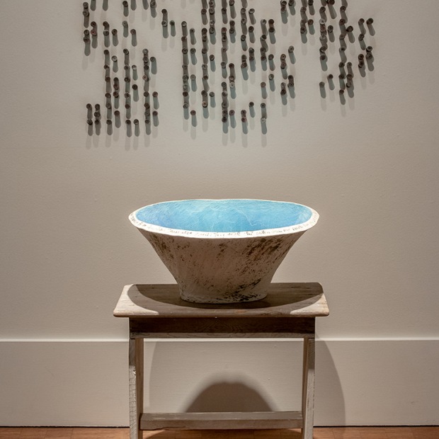A ceramic piece by Ingrid Lilligren, featuring Braille text attached to the wall behind a small wooden table with a ceramic bowl. The inside of the bowl is sky blue.
