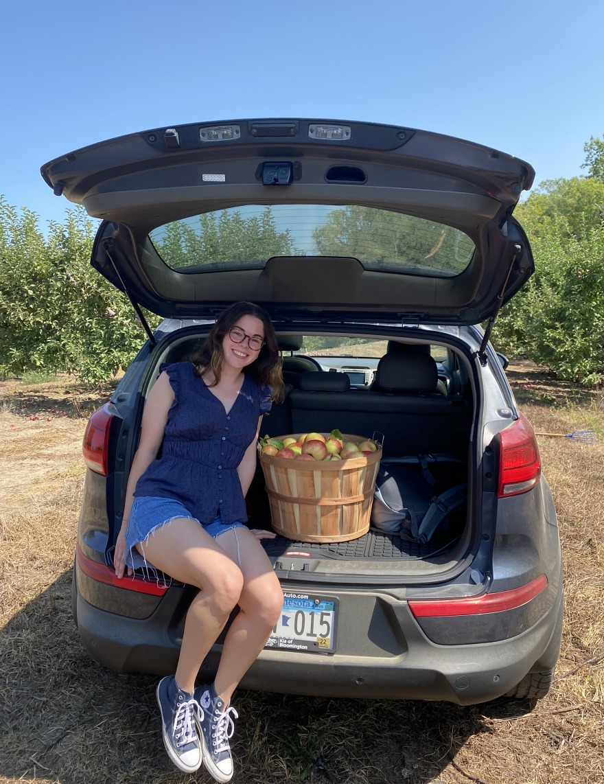 Hannah sits next to a basket of apples on the tailgate of a car