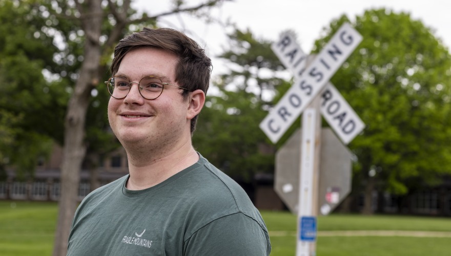 A man with short brown hair and glasses smiles in front of the railroad crossing sign.