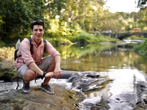 wearing a backpack, Andrew crouches on a stone in a shallow stream with grassy banks and a stone bridge in the background