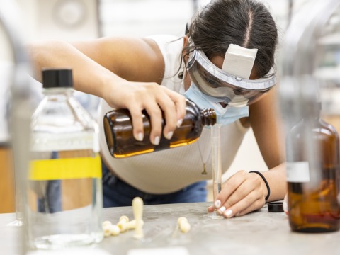 A girl with laboratory goggles leans over a bench, carefully pouring a reagent from a brown bottle into a graduated cylinder.