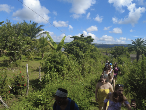 A line of students trek uphill through lush foliage in the Brazilian countryside. The sky is blue and dotted with clouds.