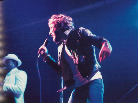 Bruce Springsteen sings into a microphone onstage at Grinnell with Clarence Clemons playing a saxophone in the background