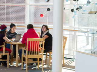 four students sit at a circular table near a large sunny window. A model of a molecule hangs above them