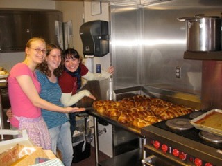 Three women in an industrial style kitchen stand to the left of and gesture toward a tray of baked challah loaves