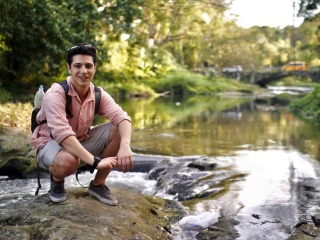 wearing a backpack, Andrew crouches on a stone in a shallow stream with grassy banks and a stone bridge in the background