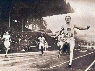 A young man in team USA uniform runs, arms out to the side, towards the finish line. Other racers follow several feet behind