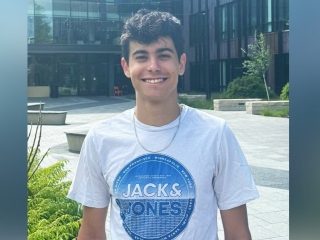 Mikel de Julian, a young man with black hair, smiles in front of a building. He wears a white shirt with a blue logo.