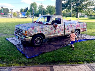 A parked truck with shiny, sparkly sequins sitting on a park lawn. A child run towards it.