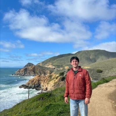 Alex Stein stands on a path near the sea. Low cliffs topped with grass and dropping off to the water are visible behind him.