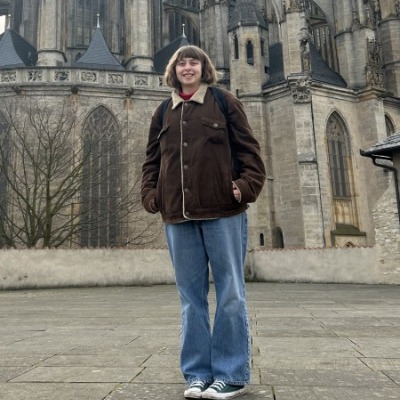 Nina Bigelow stand in front of a gothic cathedral wearing a brown jacket and blue jeans